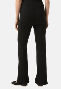 Schwarze Schlaghosen aus weichem Stoff, mit hohem Bund und glatter Textur. Kombiniert mit schwarzen Slippern.