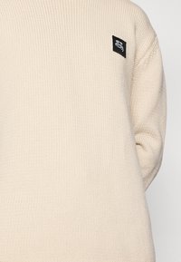 Pull en laine de couleur crème avec un design à côtes texturé et un patch logo noir sur l'épaule gauche.
