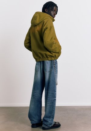 Moški z dreadlocksi, oblečen v maslinasto zeleno jopico s kapuco, široke modre kavbojke z verigo in črne čevlje, stoji na betonski tlakovani površini ob belem zidu.