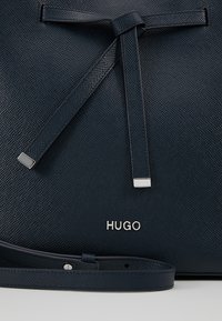 Gros plan d'un sac à main bleu foncé texturé avec une sangle nouée et des détails en métal argenté, portant le nom de la marque HUGO en lettres argentées.