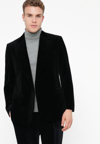 Giovane uomo che indossa un blazer doppio petto di velluto nero sopra un maglione a collo alto grigio chiaro, in piedi contro uno sfondo bianco.