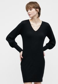 Žena s krátkymi vlasmi, oblečená v čiernych dlhých rukávoch s výstrihom do V, stojí s rukami na bokoch proti jednoduchému pozadiu.