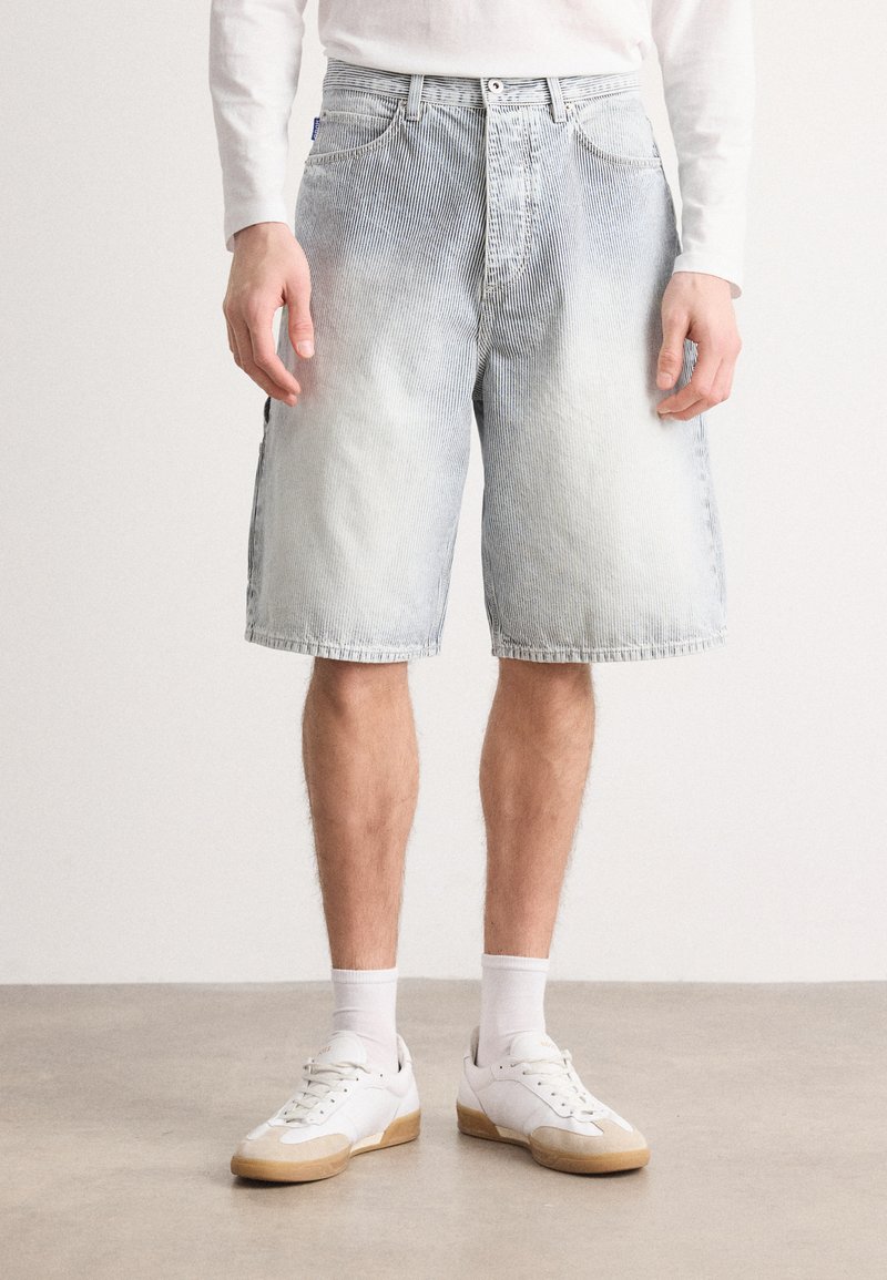 Shorts din denim de culoare albastru deschis, cu spălare uzată, croială dreaptă, buzunare frontale și talie, asortați cu adidași albaștri și șosete tip crew.