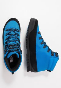 Modré turistické boty s černými semišovými detaily, texturovanou látkou, šesti kovovými průchodkami a robustní gumovou podrážkou. Logo značky uvnitř boty.