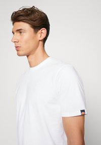 Hvid bomulds t-shirt med rund halsudskæring, der har et diskret logo på brystet og en farvet detalje på ærmet. Korte ærmer.