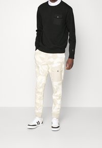 Svart långärmad sweatshirt med bröstficka, kombinerad med beige camouflagemönstrade sweatpants med sidofickor och elastiska muddar, bärs med vita sneakers.