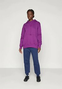 Fialová mikina s predným vreckom a kapucňou so sťahovacou šnúrkou, skombinovaná s námorníckymi nohavicami a čiernymi športovými topánkami. Hladká textura, voľný strih.