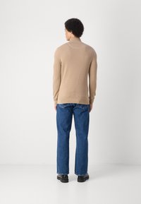 Maglione in maglia beige, aderente, con colletto alto, indossato con jeans blu a gamba dritta e scarpe nere. La texture appare liscia, con dettagli minimi.