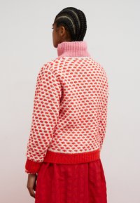 Strickpullover in Rot und Weiß mit einem rosa Kragen; mit einem strukturierten Muster und einem gerippten Saum. Wird über einem mehrlagigen roten Rock getragen.