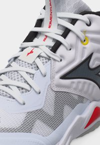 Λευκό και γκρι αθλητικό παπούτσι με ύφασμα δίχτυ, λευκά κορδόνια, μαύρες και κίτρινες λεπτομέρειες, και κόκκινο λογότυπο στη γλώσσα και στη σόλα.