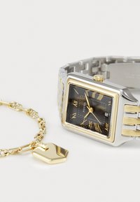 Montre Fossil carrée avec un cadran noir, des chiffres romains en or, un bracelet en métal argenté et doré. Comprend un charme doré sur un bracelet lié.