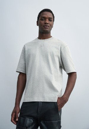 Jeune homme portant un T-shirt gris clair à manches courtes et un pantalon cargo noir, debout avec une main dans la poche devant un fond uni.