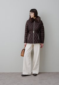 Casaco acolchoado castanho com gola de pelo sintético, cintura com cinto, fechos de correr prateados, combinado com calças largas brancas e uma mala castanha.