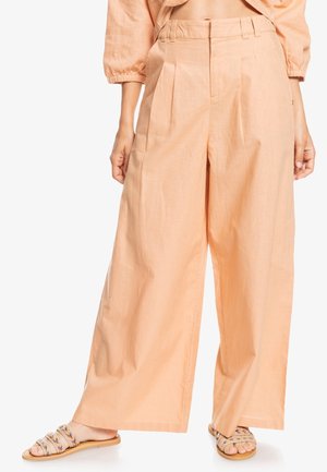 Pantalon ample couleur pêche avec plis, associé à des sandales plates beiges avec des brides tissées multicolores.