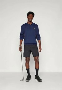 Marineblå poloshorte med gul logo, kombinert med svarte shorts. Modellen holder en golfkølle og har på seg svarte sko og sokker.