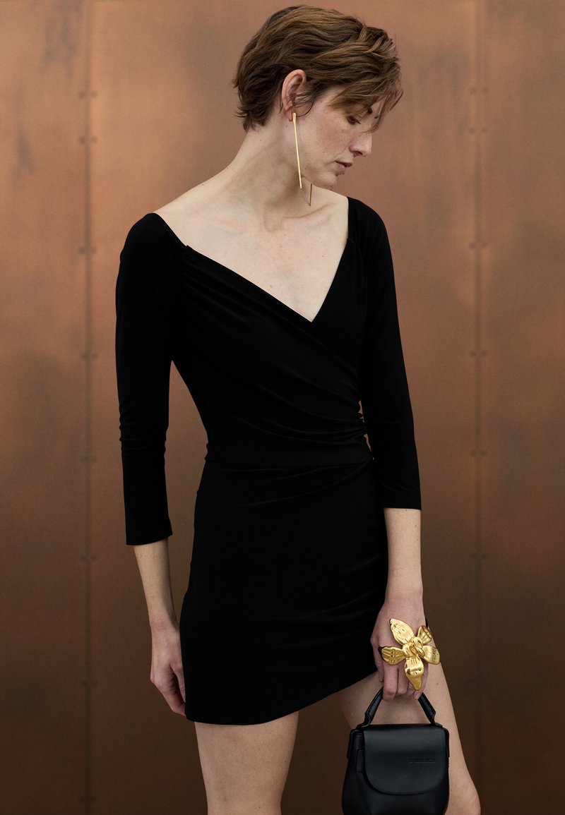 Černé zavinovací šaty s dlouhými rukávy, výstřihem do V a texturovanou látkou. Modelka drží malou černou kabelku a nosí zlatý květinový prsten.