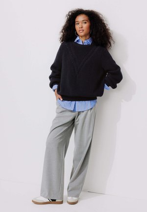 Pull en tricot noir avec un design en forme de V, superposé à une chemise à rayures bleues et blanches, associé à un pantalon large gris clair et des baskets.