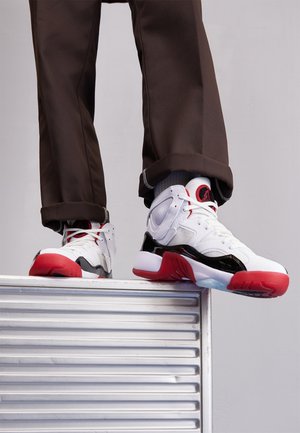 Personne portant des baskets blanches, noires et rouges, debout au bord d'une plateforme métallique striée sur un fond clair uni.