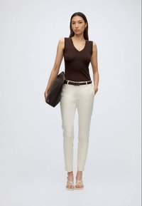 Frau trägt ein ärmelloses dunkelbraunes Top, hellbeige Hose, weiße Riemchensandalen, hält eine große dunkelbraune Tasche und steht vor einem schlichten Hintergrund.