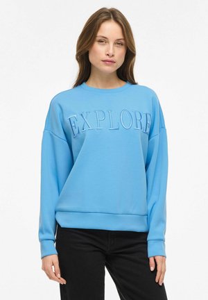 Kvinde iført lyseblå sweatshirt med teksten "EXPLORE" over brystet og sorte bukser, stående mod en ensfarvet hvid baggrund.