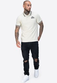 Cremfarbenes Poloshirt mit schwarzen Streifen am Kragen und an den Ärmeln, Logo auf der Brust, kombiniert mit zerrissenen schwarzen Jeans und weißen Sneakers.
