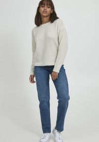 Pull en tricot crème avec un col rond et un ourlet côtelé, associé à un jean bleu et des baskets blanches. Texture douce, coupe décontractée.