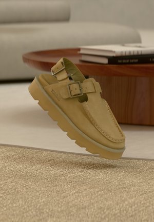 Sandalia deslizante de cuero color tan con doble hebilla, suela gruesa de goma y costuras visibles sobre una alfombra beige texturizada cerca de una mesa de madera.