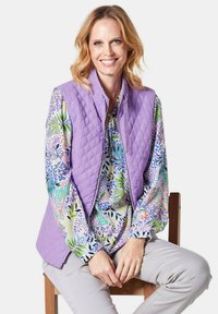 Helllila gesteppte Weste mit Reißverschluss über einer bunten Bluse mit floralen Motiven in Grün, Blau und Pink, kombiniert mit hellgrauen Hosen.