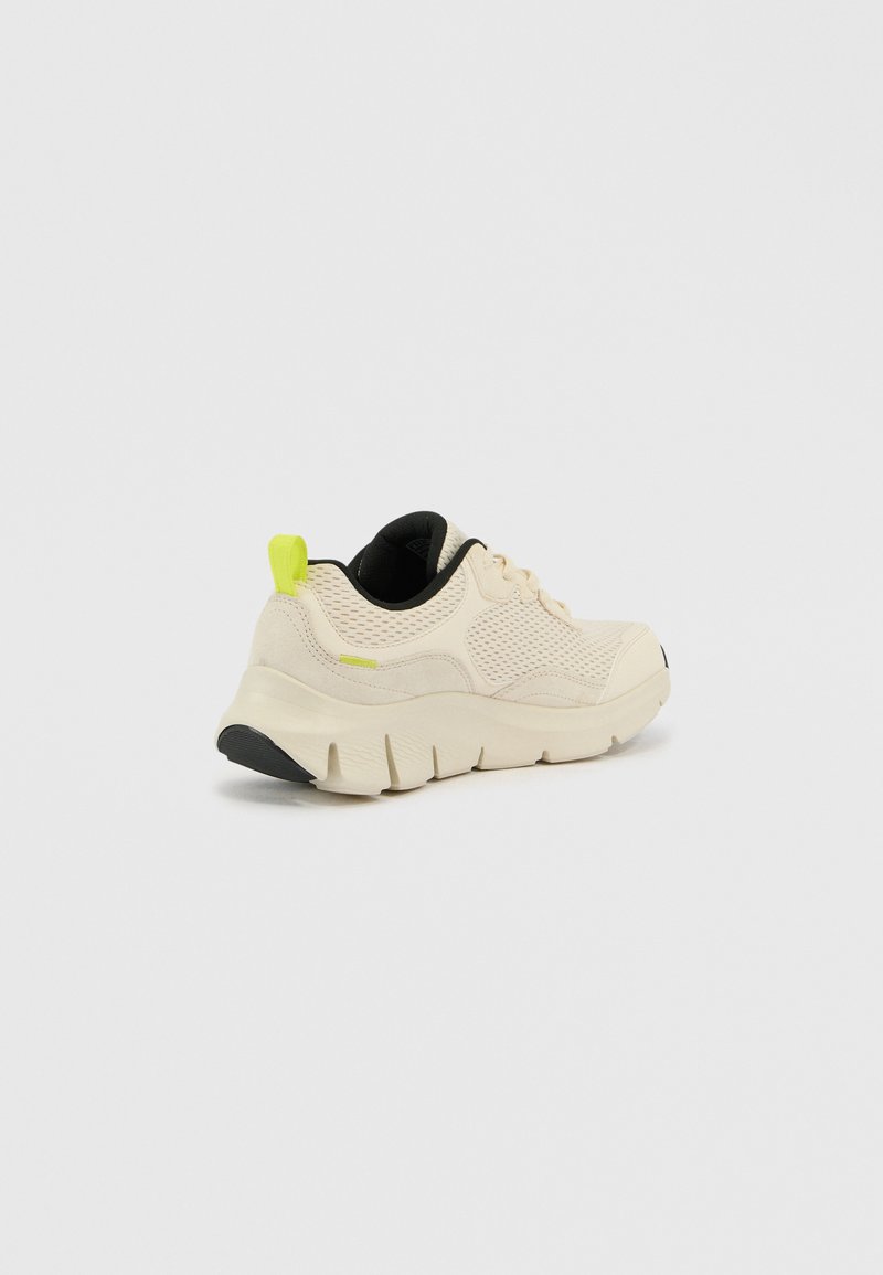 Zapatilla deportiva beige con parte superior de malla, forro interior negro y acento en verde neón en el talón. Suela blanca gruesa con suela exterior negra texturizada.