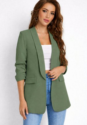 Femme aux longs cheveux ondulés portant un blazer vert sur un crop top blanc et un jean bleu, posant sur un fond clair uni.