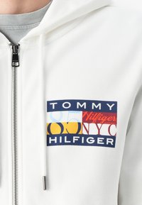 Biela mikina na zips s farebným grafickým logom a modrými, žltými, červenými a námorníckymi akcentmi. Hladká látka s voľným strihom.