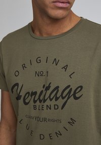 Maslinasto zelena pamučna majica s okruglim izrezom, s crnim grafičkim tekstom: "Heritage Blend" i "Original No.1" u podebljanim fontovima.