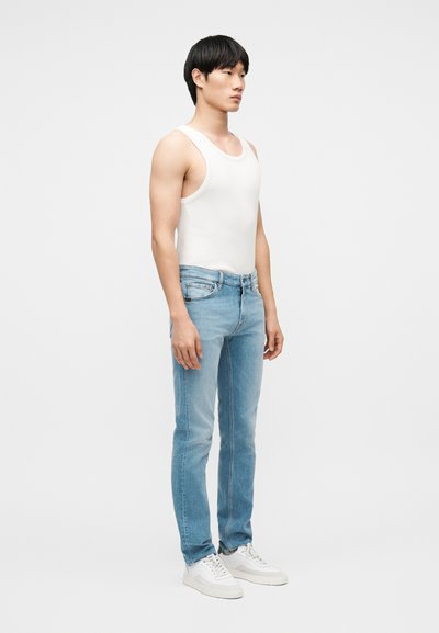 Νεαρός άνδρας που στέκεται πλαγίως, φορώντας λευκή αμάνικη μπλούζα, ανοιχτόχρωμα μπλε τζιν και λευκά αθλητικά παπούτσια, μπροστά σε απλό φόντο.