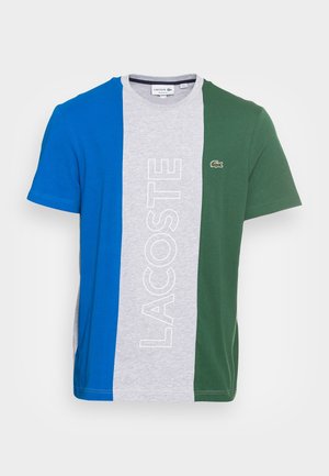 Lacoste T-shirt med korte ærmer i lodrette blå, grå og grønne farveblokke, stor hvid "LACOSTE"-tekst og lille krokodillelogo på det grønne panel.