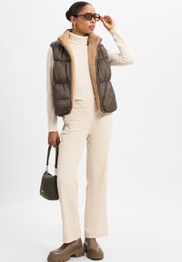 Dunkelbraune Pufferweste mit einem beigen Fleecekragen, kombiniert mit strukturierten cremefarbenen Hose und klobigen braunen Stiefeln. Grünes Handtaschenakzent.