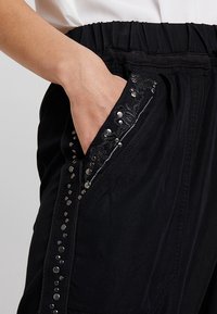 Ręka w kieszeni czarnych spodni z elastycznym paskiem, z dekoracyjnym szyciem i srebrnymi ćwiekami wzdłuż krawędzi kieszeni.