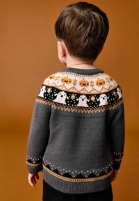 Jeune enfant vu de dos, portant un pull gris avec des citrouilles orange et des motifs de fantômes blancs sur le yoke, sur un fond marron.