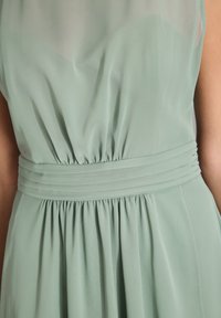 Abito verde chiaro, fluido, con vita arricciata. Realizzato in tessuto morbido, presenta pieghe sotto la vita e uno strato superiore leggermente trasparente.