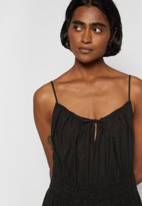 Vestido preto sem mangas com decote franzido, frente em forma de chave e alças ajustáveis. Possui uma cintura elástica para um ajuste descontraído.