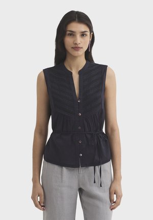 Woman wearing sleeveless black button-up blouse with textured front and waist tie, paired with light gray trousers, standing against plain background.
