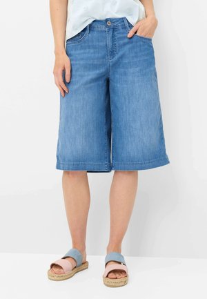 Frau trägt knielange blaue Jeansshorts und zweifarbige rosa-blaue Slip-On-Sandalen, steht vor einem schlichten weißen Hintergrund.