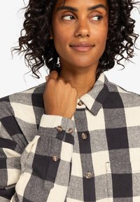 Black and white checkered shirt with buttons, made from a textured fabric. Features a collar and cuffed sleeves with decorative stitching.