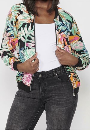 Femme portant une veste zippée fleurie colorée sur un haut blanc et un jean noir, tenant la fermeture éclair de la veste avec les deux mains.