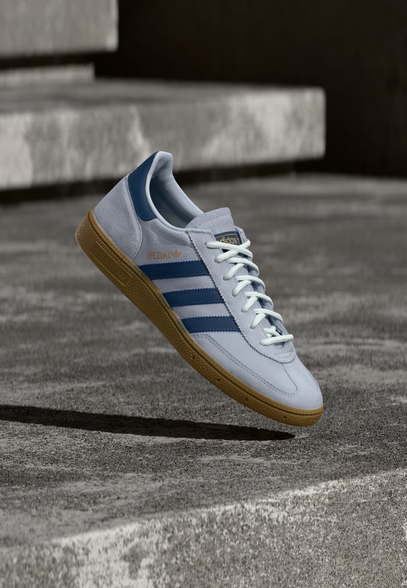Baskets Adidas Spezial bleu clair avec des rayures bleu marine et une semelle en gomme, montrées en l'air au-dessus d'escaliers en béton dans un environnement extérieur neutre.
