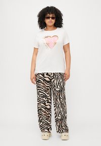 Tricou alb cu o inimă roz și accente aurii, asortat cu pantaloni bej largi cu dungi de zebră negre și maro și saboți.