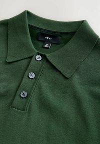 Zöld póló, texturált anyagból, lapos gallérral, három sötét gombbal és egy "NEXT" címkével, amely a karbantartási részleteket tartalmazza.