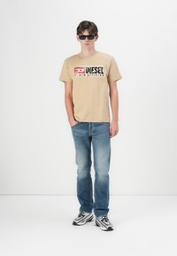 T-shirt en coton beige avec impression graphique, associé à un jean en denim bleu et des baskets noir et blanc. Le modèle porte des lunettes de soleil sombres.