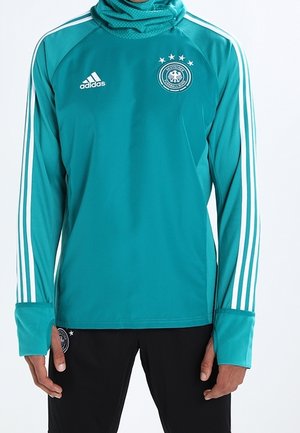 Chemise de sport à manches longues teal avec col haut, présentant trois bandes blanches sur les manches et un emblème circulaire allemand sur la poitrine.