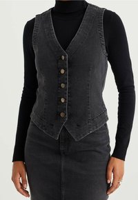 Svart denimväst med V-ringning, fem metallknappar och strukturerade sömmar; bärs över en långärmad svart polotröja.