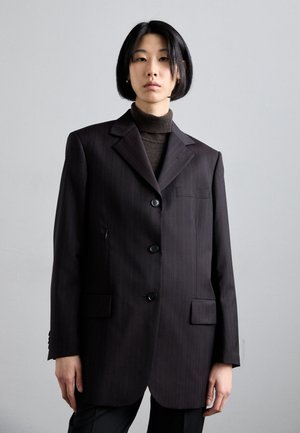 Blazer a righe scure con rever a punta, tre bottoni, due tasche frontali e vestibilità su misura. Indossato su un maglione a collo alto scuro.